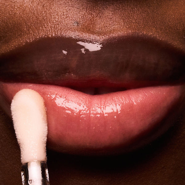 A close-up of glossy lips as a doe-foot applicator applies a clear, shiny lip serum to the lower lip. The product creates a smooth, hydrated finish with a high-shine look.