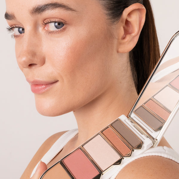 A woman with natural, glowing makeup poses with an open makeup palette resting on her shoulder. The palette contains warm neutral shades, including pink, champagne, bronze, and brown tones, shown both directly and reflected in its built-in mirror.