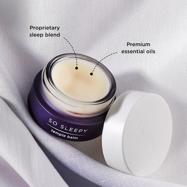 Open jar of Tropic So Sleepy Temple Balm resting on soft white fabric, showing creamy balm texture with labels pointing to its proprietary sleep blend and premium essential oils.