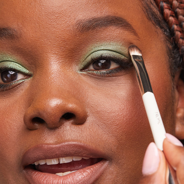 A close-up of a person applying shimmery green eyeshadow using a flat detailing makeup brush near the outer corner of their eye.