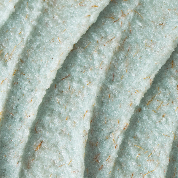 A close-up texture shot of Foot Smooth Enzymatic Scrub, showing its pale mint-green formula spread in soft, sculpted ridges. The scrub has a creamy yet grainy consistency, with visible natural exfoliating particles dispersed throughout, creating a gently buffing, skin-polishing texture.