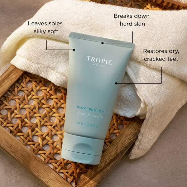 Tropic Foot Remedy softening buttercream on a wicker tray with a towel, with callouts highlighting leaves soles silky soft, breaks down hard skin and restores dry cracked feet.