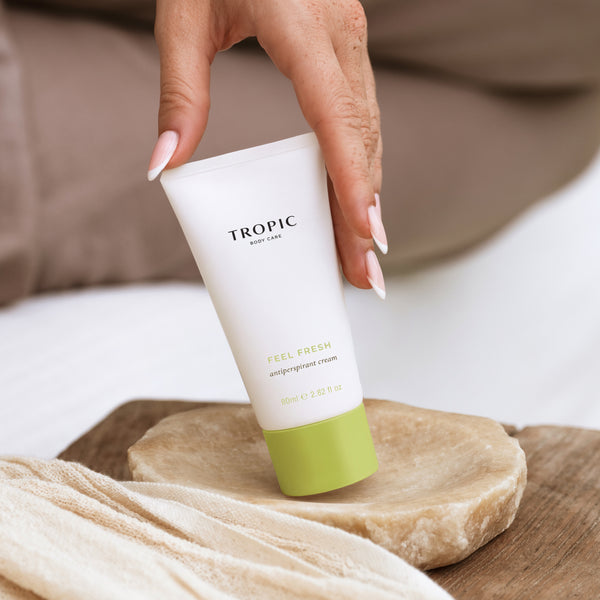 A hand with pale pink manicured nails holds a white tube of Tropic Feel Fresh Antiperspirant Cream, which has a light green cap, resting on a stone dish over a wooden surface.