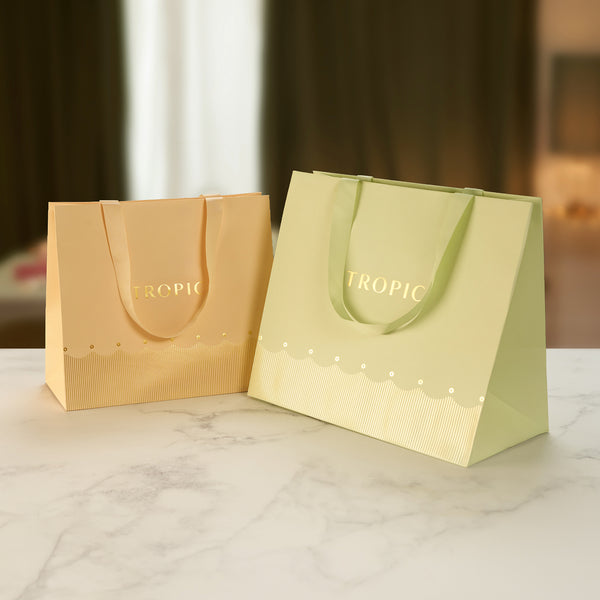 Two Tropic branded gift bags in gold and sage green with gold foil logo on a marble surface.