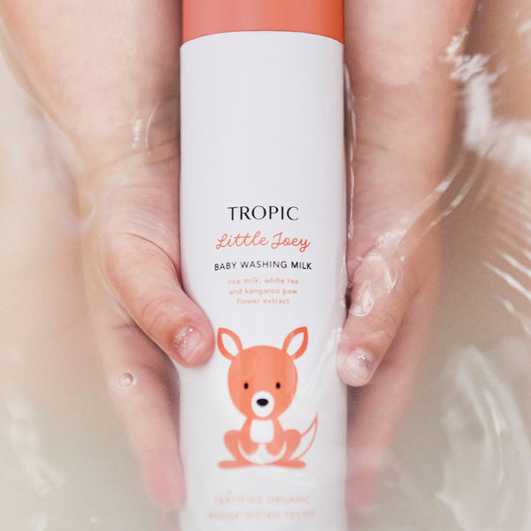 Hands holding a Tropic Little Joey Baby Washing Milk bottle with an orange cap and kangaroo illustration, partially submerged in water.