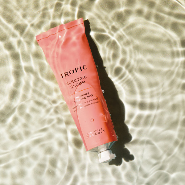 This image features a tube of Electric Bloom, a brightening tightening mask. The coral pink packaging highlights its vibrant and rejuvenating properties. Key ingredients include agave nectar, electric daisy, and sea buckthorn extracts. The product is certified organic, and the rippling water backdrop adds a refreshing and invigorating aesthetic.