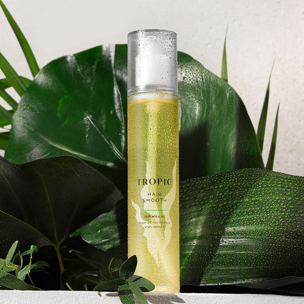 Tropic Hair Smooth Radiance Oil bottle with a clear cap, covered in water droplets, surrounded by lush green tropical leaves, creating a fresh, natural setting.
