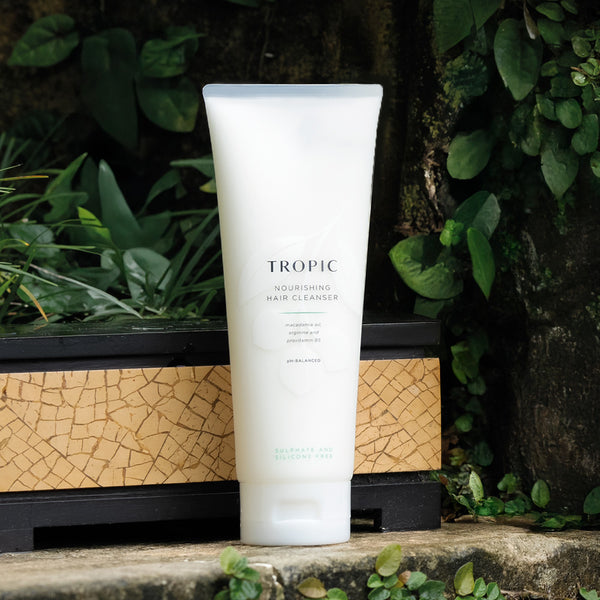 Tropic Nourishing Hair Cleanser tube placed outdoors on a natural stone surface surrounded by lush green foliage, emphasising its plant-based formulation.