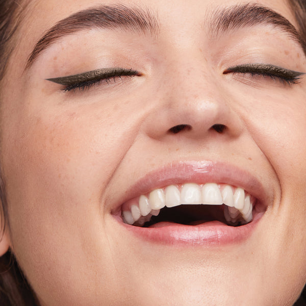 A close-up of a smiling face with natural skin, glossy lips, and winged eyeliner.