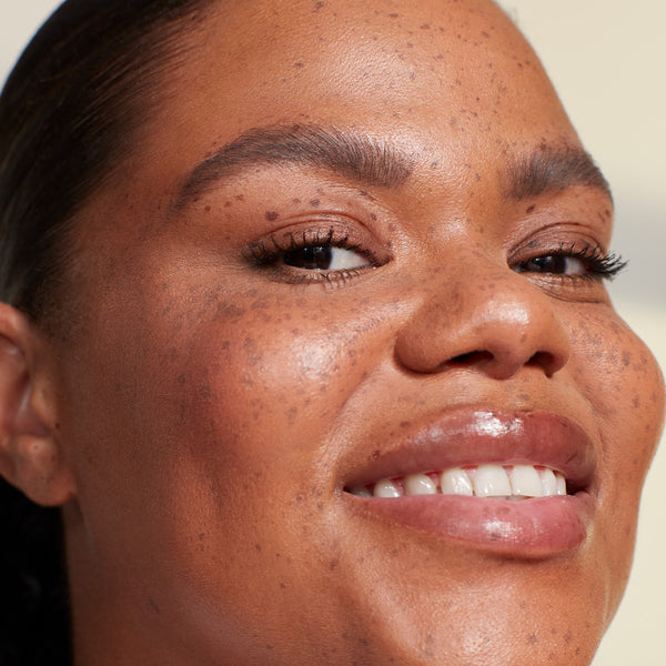 A close-up of a smiling woman with freckles, wearing natural makeup with a soft glow, defined lashes, and glossy lips.