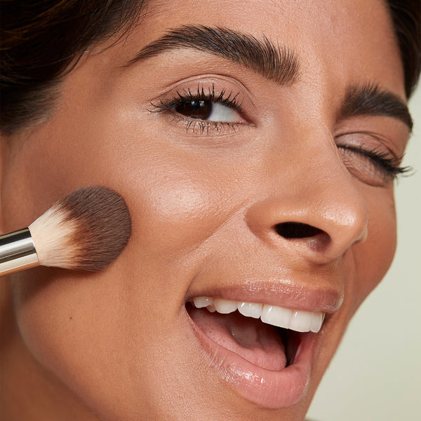 A close-up of a woman playfully smiling and winking while applying makeup to her cheek with a fluffy powder brush.