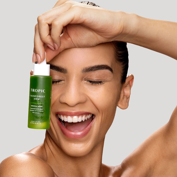 Laughing woman holding Tropic Rainforest Dew hydration serum beside her glowing face against a grey background.