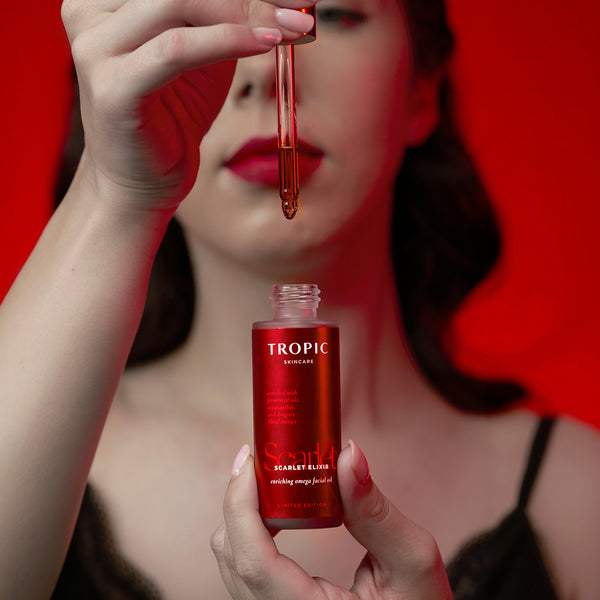 A woman holds a bottle of Tropic Skincare Scarlet Elixir omega facial oil and a glass dropper filled with the amber-red liquid, set against a solid red background.