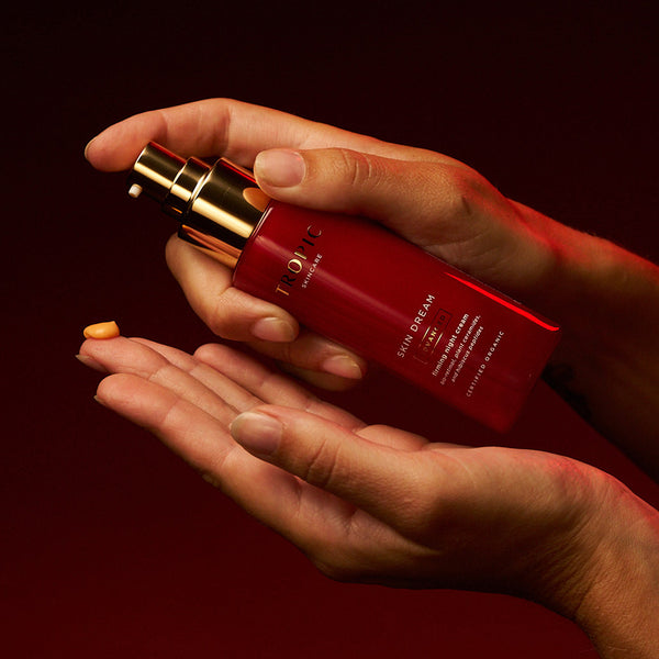 Hands dispensing a pump of Tropic Skincare Skin Dream Advanced Firming Night Cream, with a drop of peach cream visible on the palm.