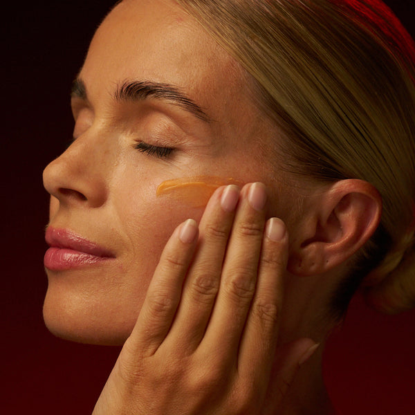 Woman massaging peach cream onto her cheekbone with eyes closed, against a deep red background.