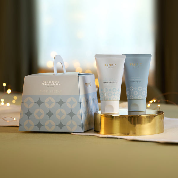 Tropic Coconut & Tonka Macaron exfoliating body cleanser and nourishing body cream on a gold tray beside the patterned seasonal gift bag with fairy lights.