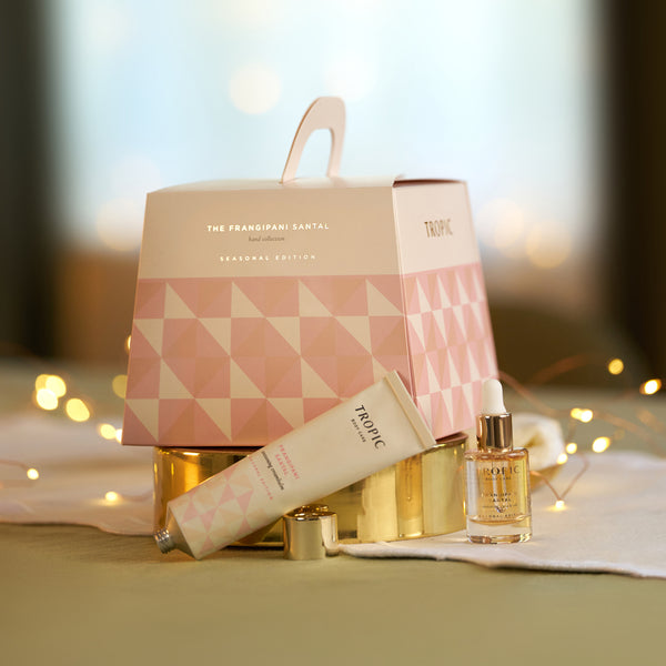 Tropic Frangipani Santal hand cream and restoring face oil beside the pink geometric seasonal gift bag with fairy lights.