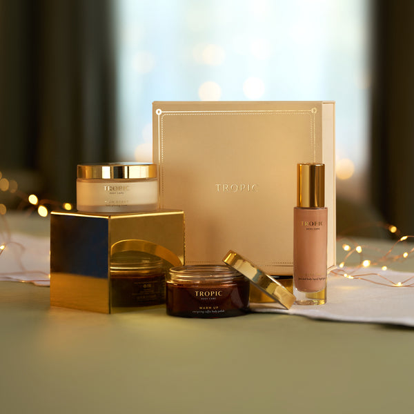 Tropic gift set with Main Event Body Glossing Cream, Warm Up Coffee Body Polish and All Eyes On You Liquid Highlighter beside gold gift box with fairy lights.