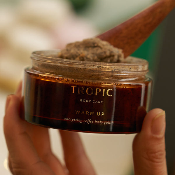 Hands holding open jar of Tropic Warm Up Energising Coffee Body Polish with wooden spatula.
