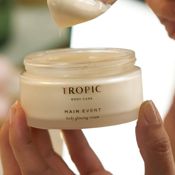 Hands scooping Tropic Main Event Body Glossing Cream from open jar.