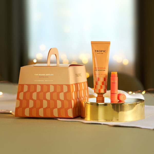 Tropic Mango Brûlée nourishing hand cream and lip balm on a gold tray beside the orange geometric seasonal gift bag with fairy lights.