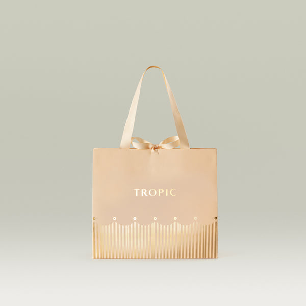Tropic gift bag in blush and gold with a satin ribbon handle and bow, against a neutral background.