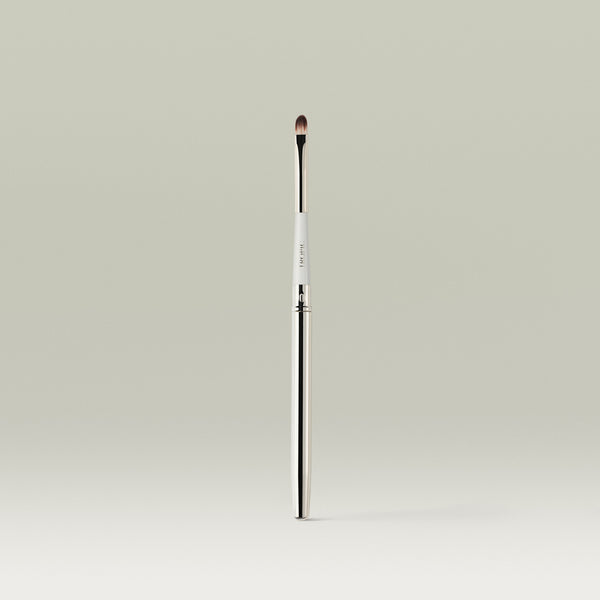 A sleek, dual-ended lip brush with a slim metallic handle and a small, rounded bristle tip, standing upright against a soft neutral background.