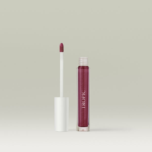 Open tube of Tropic Lip Glaze with the applicator wand standing beside the transparent gloss bottle against a neutral background. Shade Sugar Plum.