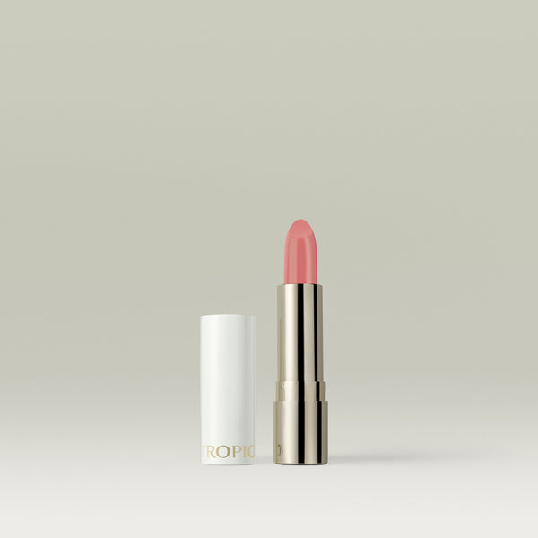A soft peach-pink lipstick in a metallic silver tube, shown uncapped with the white lid placed beside it against a soft, neutral background.