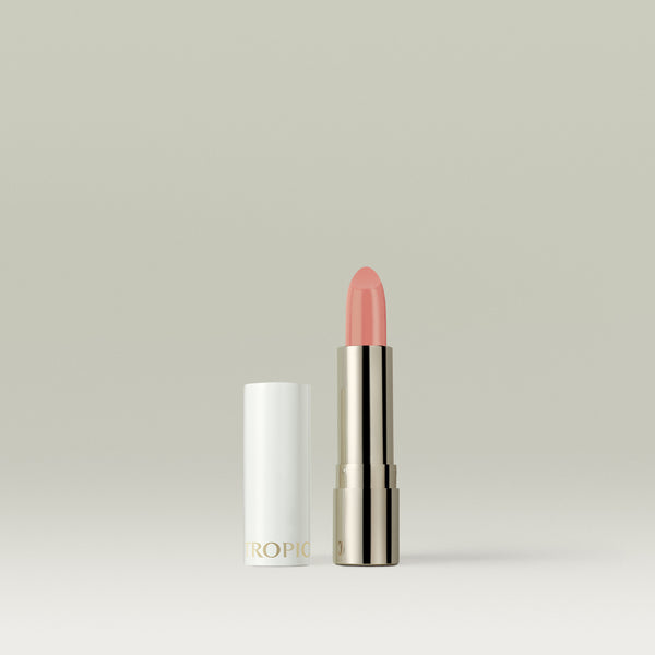 A soft peachy-pink lipstick in a metallic silver tube, shown uncapped with the white lid placed beside it against a soft, neutral background.