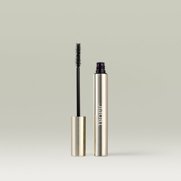 Silver tube of Tropic mascara  in shade black with the wand removed and standing beside it, showing a black bristle brush coated in product. 