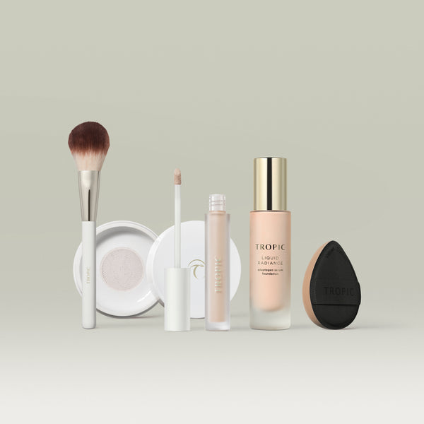 A row of Tropic makeup products in shade 1 displayed on a soft neutral background, including a fluffy makeup brush, a white compact powder case, a  concealer with a doe-foot applicator, a bottle of Liquid Radiance foundation with a gold cap, and a black teardrop-shaped blending sponge.