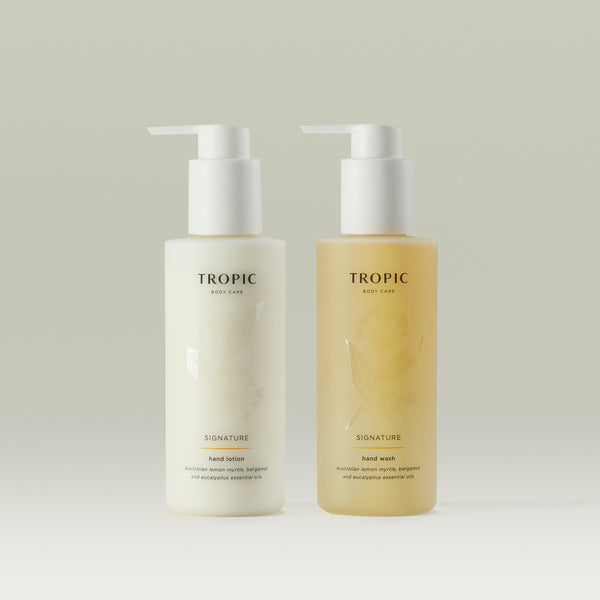 Two bottles of Tropic Body Care Signature hand care products, featuring a hand lotion and a hand wash. Both bottles have a minimalist design with white pump dispensers and subtle floral patterns on the semi-transparent packaging. The hand lotion is in a white bottle, while the hand wash is in a light amber-colored bottle. The labels display 'TROPIC BODY CARE' at the top, followed by 'SIGNATURE' and product details, mentioning Australian lemon myrtle, bergamot, and eucalyptus essential oils.