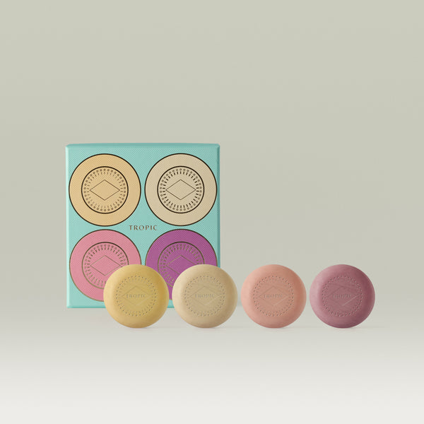Four Tropic embossed body pebbles in yellow, cream, peach, and mauve, displayed in front of a teal gift box featuring a bold four-circle graphic design.