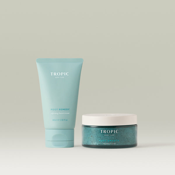 Two Tropic Body Care foot-care products on a clean, light beige background: a pastel turquoise tube labeled “Foot Remedy” softening buttercream standing beside a matching jar labelled “Foot Smooth” enzymatic scrub with a white lid.