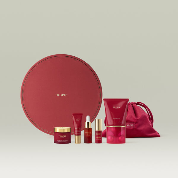 Tropic luxury skincare gift set in deep red, including Renewing Cleanser, Skin Dream age-defying night cream, Youth Potion, Lip Silk, and Neck Sculpt lifting collagen cream, with a matching satin drawstring pouch and large circular gift box.