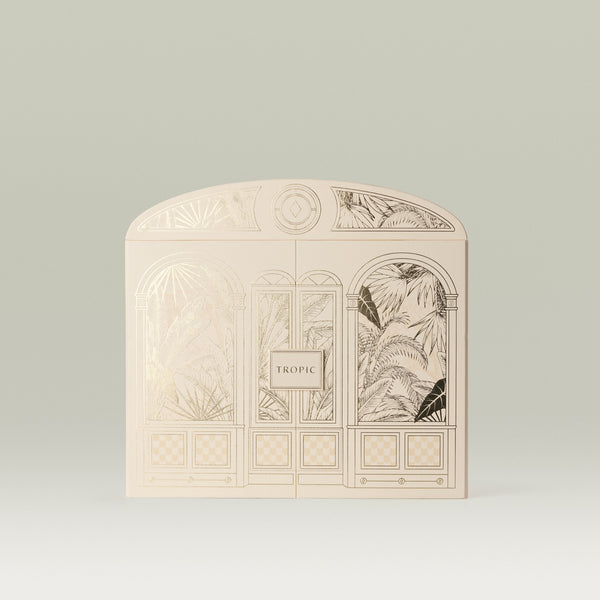 Tropic arch-shaped gift box in cream, featuring an illustrated building facade with gold foil tropical palm leaf detailing, arched windows, and a checkerboard base panel.